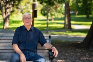 Older man sitting on a park bench outdoors, smiling, representing a patient whose life was saved by early heart screening.
